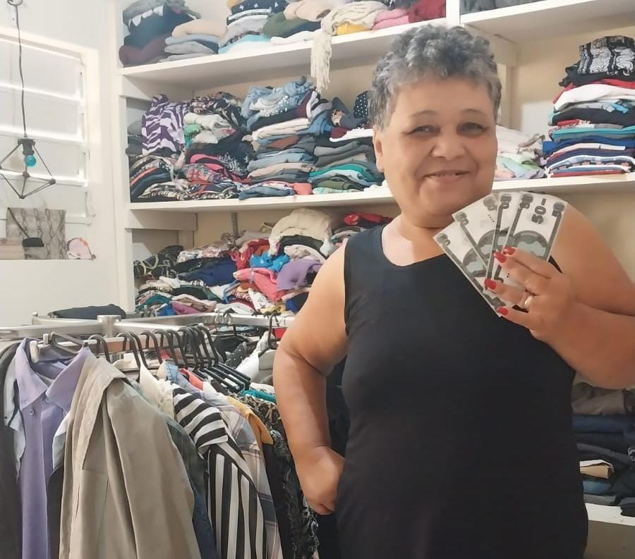 Na imagem, uma senhora segura notas de "Reais CDI", e sorri para a câmera, ela veste blusa preta e suas unhas estão pintadas de vermelho. Ao fundo podemos ver a lojinha do local, com diversas roupas em estantes ao fundo, e em cabides mais a frente.